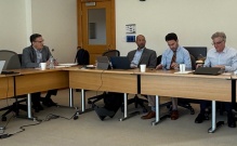 Four people are seated around a U-shaped conference table in a meeting room. The table holds laptops, papers, coffee cups, and water bottles. 