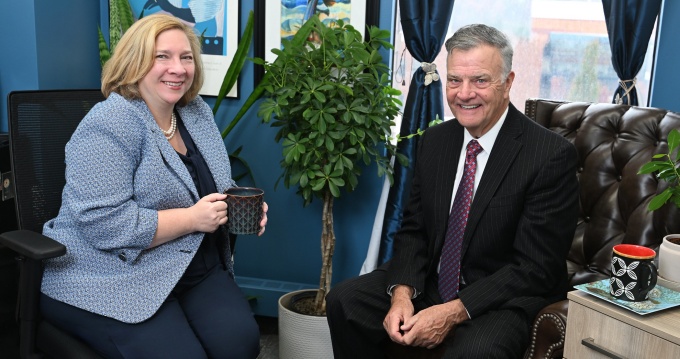 Two people are seated in an office setting, engaged in conversation. One person is holding a black coffee mug while sitting on an office chair, and the other is seated on a tufted leather chair. The background includes framed artwork on blue walls and windows with dark curtains. 