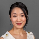 A person wearing a sleeveless white top with gold button details on the shoulders is posed against a plain gray background. 