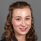 A person with shoulder-length curly hair is wearing a dark blazer over a teal top, posed against a plain gray background in a professional portrait style. 