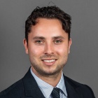 A person wearing a dark suit jacket, a light blue striped dress shirt, and a dark patterned tie is posed against a plain gray background in a professional headshot style. 