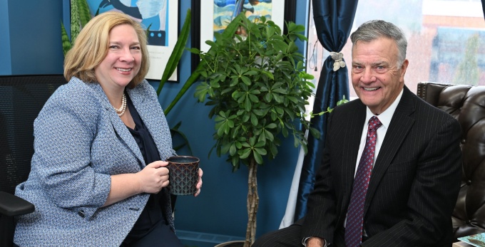 Two people are seated in an office setting, engaged in conversation. One person is holding a black coffee mug while sitting on an office chair, and the other is seated on a tufted leather chair. The background includes framed artwork on blue walls and windows with dark curtains. 