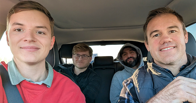 Four people seated inside a car, two in the front seats and two in the back, with seat belts visible and sunlight coming through the windows. 