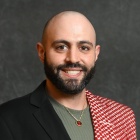 Person wearing a black blazer over a textured green shirt, accessorized with a gold necklace featuring a red pendant. A red and white patterned scarf is draped over one shoulder. The background is a plain dark gray backdrop. 