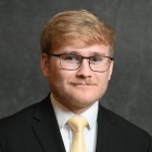 Person wearing a black suit jacket over a white dress shirt, paired with a solid light yellow tie. The background is a plain dark gray backdrop. 