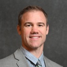 Person wearing a light gray suit jacket over a light blue dress shirt, paired with a patterned tie featuring diagonal stripes in shades of green, gray, and black. The background is a plain dark gray backdrop. 