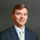 Person wearing a dark navy suit jacket over a light blue dress shirt, paired with a light-colored tie featuring a floral pattern in pink, green, and white. The background is a plain dark gray backdrop. 