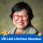 A person wearing red glasses, a gray jacket, and a beaded necklace smiles at the camera against a muted green background. A blue banner at the bottom reads "UB LAA Lifetime Member" in white text, flanked by two gold stars. 
