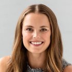 A person with long, light brown hair and a bright smile poses against a plain, light-colored background. 