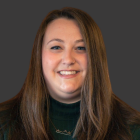 Person with long, straight brown hair wearing a dark green turtleneck sweater and a geometric gold necklace, posed against a solid dark gray background. 