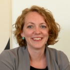 A person with short blond, wavy hair, turquois earrings, grey jacket, smiling. 