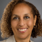 A person with curly hair, wearing hoop earrings and a light-colored blazer over a blue top, poses against a plain gray background. 