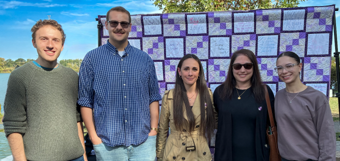 Five people stand in front of a quilt made of purple and white squares, each featuring supportive messages like “You are loved,” “Be brave,” and “Believe in yourself.” Behind them is a clear blue sky, trees, and a body of water. 