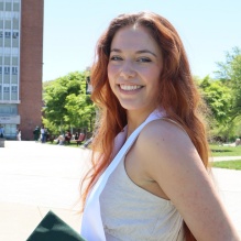 A person with long hair sits outdoors on a sunny day, holding a green graduation cap and wearing a white sash over a light-colored dress. Behind them is a tall building with many windows, trees, and people walking. 