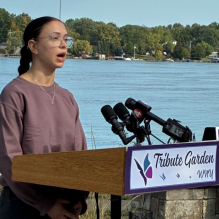 A person stands behind a podium labeled “Fairie Garden WNY” at an outdoor event near a body of water, surrounded by trees and greenery. Multiple microphones are attached to the podium. 
