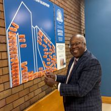 A person in a plaid suit jacket is placing an orange card on a large blue display titled 'Your Law Blueprint.' The display features an outline of buildings filled with numerous orange cards containing handwritten messages. A smaller sign on the right explains the blueprint activity and includes the phrase 'Our Blueprints' with an illustrated logo. 
