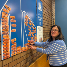 A person wearing a blue-and-white striped sweater is placing an orange card on a large blue display titled 'Your Law Blueprint.' The display features an outline of buildings filled with numerous orange cards containing handwritten messages. A smaller sign on the right explains the blueprint activity and includes the phrase 'Our Blueprints' with an illustrated logo. 