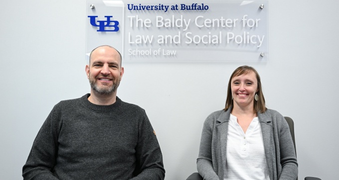 Matthew Dimick and Amanda Benzin sitting together at a table. Sign behind them says "University at Buffalo, The Baldy Center for Law and Social Policy, School of Law.". 