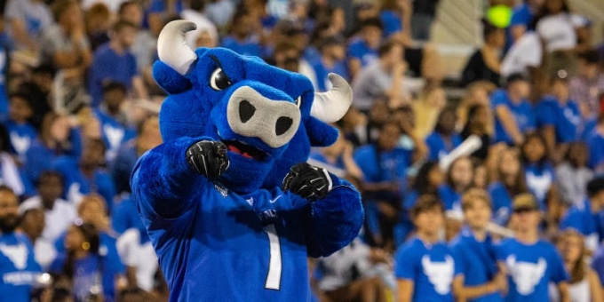 UB's mascot, Victor E. Bull, at a sporting event, crowd of attendees behind him wearing UB blue tshirts. 
