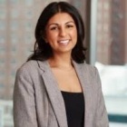 Person with short, dark hair wearing beige jacket and black top, buildings in the background. 