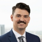 Person with mustache, wearing a blue suit jacket, striped tie, white shirt, standing in front of a window of an office building. 