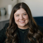 Close‑cropped professional portrait of a person with long hair, wearing a dark top and blazer, photographed indoors against a softly blurred office‑like background. 