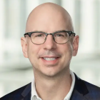Close‑cropped professional portrait of a person wearing a dark blazer over a light shirt, photographed indoors against a softly blurred, light‑colored background. 