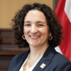 A head‑and‑shoulders professional portrait of an individual with short, curly hair, wearing a dark blazer over a light top and small hoop earrings. A red and white flag and wood‑paneled interior appear in the background. 