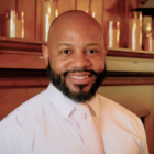 A head‑and‑shoulders professional portrait of an individual wearing a light‑colored dress shirt and a pale tie, photographed indoors against a wood‑paneled background with soft, warm lighting. 