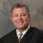 A professional head and shoulders portrait of an individual wearing a dark judicial robe over a light shirt and patterned tie, photographed against a neutral studio background. 