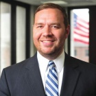 Close‑cropped professional portrait of a person wearing a dark blazer over a light shirt, photographed indoors against a softly blurred, light‑colored background. 
