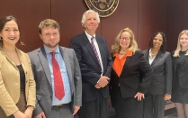 Group of seven people standing indoors in formal business attire beneath a wall mounted seal reading &ldquo;United States District Court.&rdquo; The group is positioned against a wood paneled wall in a courtroom or courthouse setting. 