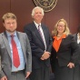 Group of seven people standing indoors in formal business attire beneath a wall mounted seal reading &ldquo;United States District Court.&rdquo; The group is positioned against a wood paneled wall in a courtroom or courthouse setting. 