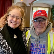 Two people standing together indoors in a casual dining or meeting space with wooden chairs and tables. One person wears a patterned jacket, and the other wears a hoodie with a safety vest. 