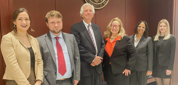 Group of seven people standing indoors in formal business attire beneath a wall mounted seal reading “United States District Court.” The group is positioned against a wood paneled wall in a courtroom or courthouse setting.. 