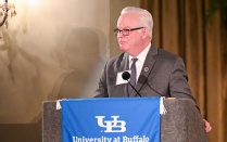 Whalen standing behind a podium and speaking into microphones at an indoor formal event. The podium is draped with a blue banner reading &ldquo;University at Buffalo School of Law.&rdquo; The setting includes warm lighting, draped curtains, and a wall mounted light fixture, suggesting a ceremonial or academic event.]. 