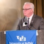 Whalen standing behind a podium and speaking into microphones at an indoor formal event. The podium is draped with a blue banner reading &ldquo;University at Buffalo School of Law.&rdquo; The setting includes warm lighting, draped curtains, and a wall mounted light fixture, suggesting a ceremonial or academic event.]. 