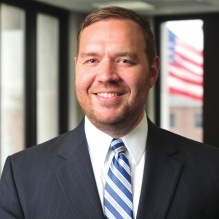 Eldridge wearing a blue suit jacket, light blue dress shirt, and pink patterned tie stands indoors in a modern building with glass walls. The background shows a softly blurred interior with natural light and architectural details, suggesting a professional or institutional setting. 