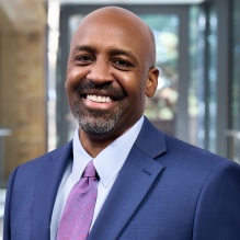 Beane wearing a blue suit jacket, light blue dress shirt, and pink patterned tie stands indoors in a modern building with glass walls. The background shows a softly blurred interior with natural light and architectural details, suggesting a professional or institutional setting. 