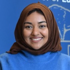Person standing indoors with hands clasped, wearing a blue sweater and dark pants. The individual stands in front of a blue wall display featuring the text &ldquo;University at Buffalo School of Law&rdquo; and a UB logo. Wood paneling and a counter are visible on either side, suggesting a professional or academic setting. 