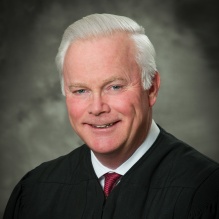 Formal studio portrait showing a person wearing a black judicial robe over a white dress shirt and red tie. The image is cropped at the shoulders, with short white hair visible, and a neutral gray background. 
