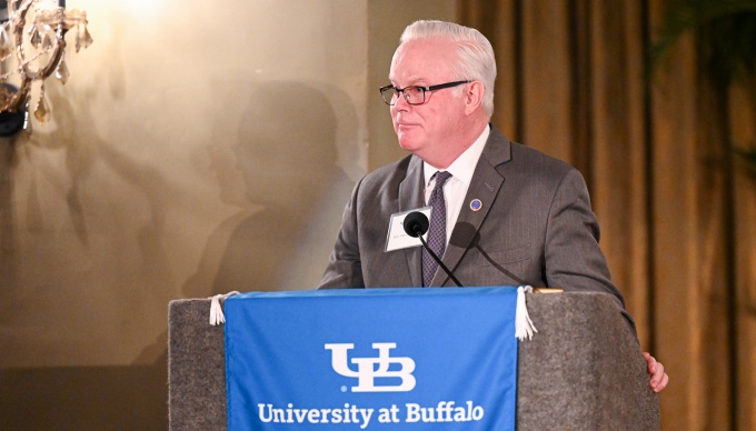 Whalen standing behind a podium and speaking into microphones at an indoor formal event. The podium is draped with a blue banner reading &ldquo;University at Buffalo School of Law.&rdquo; The setting includes warm lighting, draped curtains, and a wall mounted light fixture, suggesting a ceremonial or academic event.]. 
