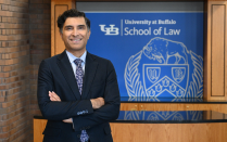 Person wearing a dark suit and patterned tie stands with arms crossed in an indoor lobby or reception area. Behind the person is a wooden desk and a blue sign displaying the text &ldquo;University at Buffalo School of Law&rdquo; along with a university crest. Brick walls and wood paneling frame the space. 