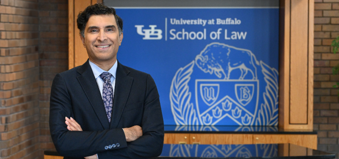 Person wearing a dark suit and patterned tie stands with arms crossed in an indoor lobby or reception area. Behind the person is a wooden desk and a blue sign displaying the text &ldquo;University at Buffalo School of Law&rdquo; along with a university crest. Brick walls and wood paneling frame the space. 