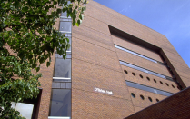 Exterior view of a multi-story brick building with narrow horizontal windows and circular window details. A vertical glass section runs along the left side, partially framed by leafy tree branches in the foreground. The building name &ldquo;O&rsquo;Brian Hall&rdquo; is visible on the brick facade under a clear blue sky. 