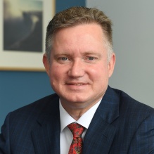 Head and shoulders portrait of Brown wearing a dark suit jacket, white dress shirt, and red patterned tie, seated indoors with a framed picture and blue wall visible in the background. 