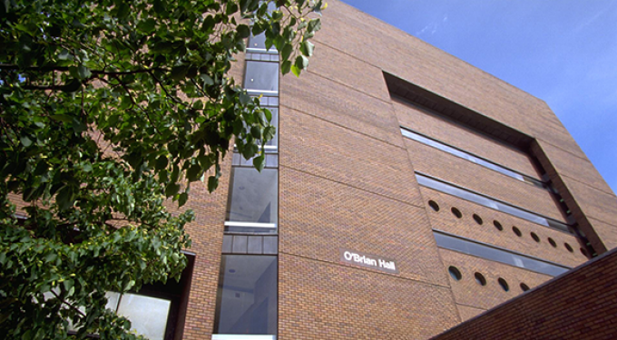 Exterior view of a multi-story brick building with narrow horizontal windows and circular window details. A vertical glass section runs along the left side, partially framed by leafy tree branches in the foreground. The building name &ldquo;O&rsquo;Brian Hall&rdquo; is visible on the brick facade under a clear blue sky. 