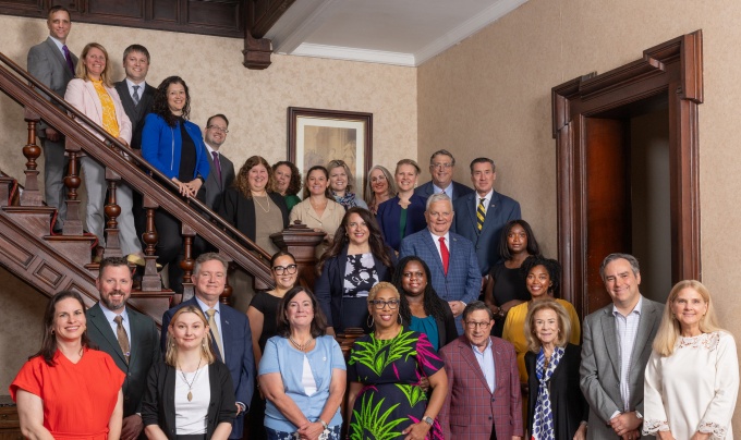 Group portrait of approximately 30 people posed on and around a grand wooden staircase inside a historic building with dark wood paneling and patterned carpeting.