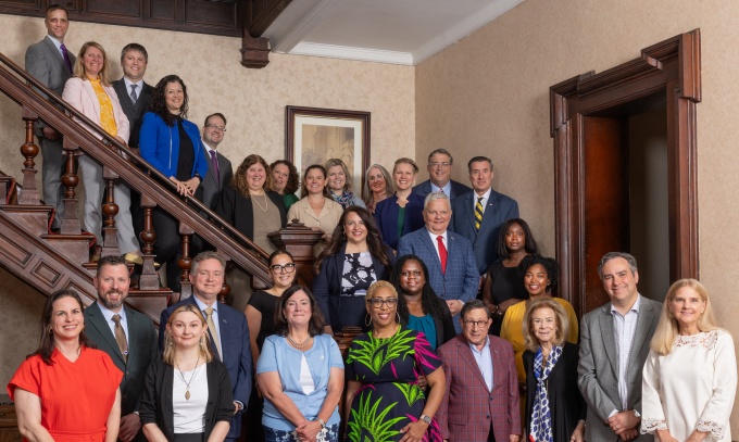Group portrait of approximately 30 people posed on and around a grand wooden staircase inside a historic building with dark wood paneling and patterned carpeting.