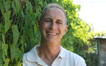 Head-and-shoulders portrait of Scott Thomas wearing a white collared shirt, photographed outdoors with green foliage and a wooden structure in the background. 